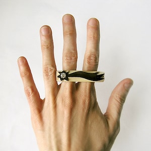 May include: A black and white ring with a star design and a clear rhinestone. The ring is worn on the middle finger of a hand.