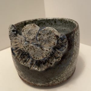 Puede incluir: Una taza de cerámica con esmalte moteado en azul y gris que presenta una flor esculpida tridimensional prominente en el exterior. El recipiente tiene una forma redondeada con una superficie texturizada y una abertura ancha, acabado rústico.