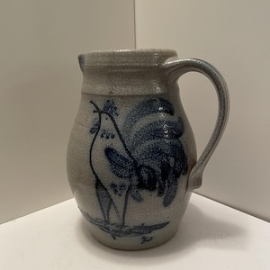 May include: A stoneware pitcher with a speckled light gray finish and a dark blue hand-painted rooster design on the front. The vessel features a curved handle on the right side, a wide body, and a small pouring spout at the top rim of the container.