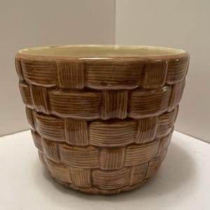 May include: A brown ceramic planter with a glossy finish and a textured exterior that mimics the appearance of a woven basket. The interior of the round vessel is a smooth, light cream colour, featuring a wide opening and a tapered base design.