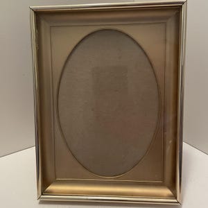 May include: A rectangular picture frame with a metallic gold finish and a polished gold-tone outer edge. The frame features a central oval-shaped matting area with a neutral beige backing, designed to hold a photograph or artwork securely.