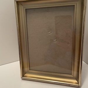 May include: A rectangular picture frame with a metallic gold finish and a beveled border. The frame features a clear glass front covering a plain, light brown cardboard backing insert that has several small circular indentations visible.