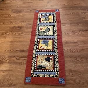 May include: A rectangular fabric table runner featuring a terracotta red border and four central square panels. Each panel displays a different rooster illustration against a cream background, framed by black and white checkered and floral fabric strips.