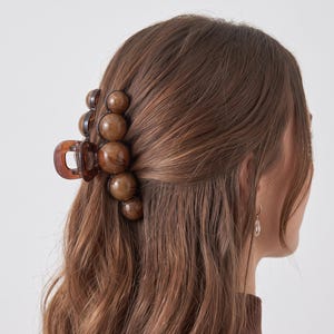 Könnte beinhalten: Eine braune Haarspange mit einer Reihe glatter, kugelförmiger Perlen mit Marmormuster entlang der Außenkante. Die Spange besteht aus einem durchscheinenden, dunkelbraunen Harzmaterial mit einem Federmechanismus, um das Haar sicher zu halten.