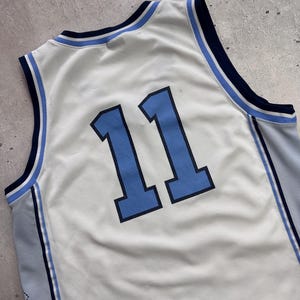 Op de afbeelding: Een mouwloos basketbalshirt van witte mesh-stof met het nummer 11 in lichtblauw met een donkerblauwe omlijning. De kraag en armsgaten hebben gestreepte biezen in donkerblauw, lichtblauw en wit, met lichtblauwe zijpanelen.