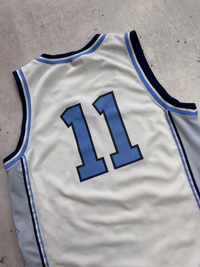 Op de afbeelding: Een mouwloos basketbalshirt van witte mesh-stof met het nummer 11 in lichtblauw met een donkerblauwe omlijning. De kraag en armsgaten hebben gestreepte biezen in donkerblauw, lichtblauw en wit, met lichtblauwe zijpanelen.