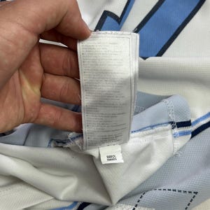 Op de afbeelding: Een wit en lichtblauw sportshirt met een donkerblauw ruitpatroon en een lang, wit waslabel aan de binnenzool. Het label bevat onderaan een kleiner wit label met de gedrukte tekst S680L 106925.