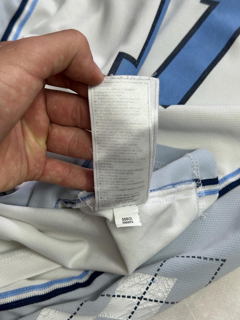 Op de afbeelding: Een wit en lichtblauw sportshirt met een donkerblauw ruitpatroon en een lang, wit waslabel aan de binnenzool. Het label bevat onderaan een kleiner wit label met de gedrukte tekst S680L 106925.