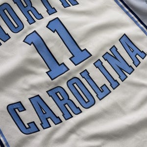 Op de afbeelding: Een lichtgrijs basketbalshirt met de tekst NORTH CAROLINA in blauwe letters met donkerblauwe omlijningen. Het nummer 11 staat in het midden gedrukt in hetzelfde blauwe en donkerblauwe kleurenschema. Een klein North Carolina logo zit op de kraag.