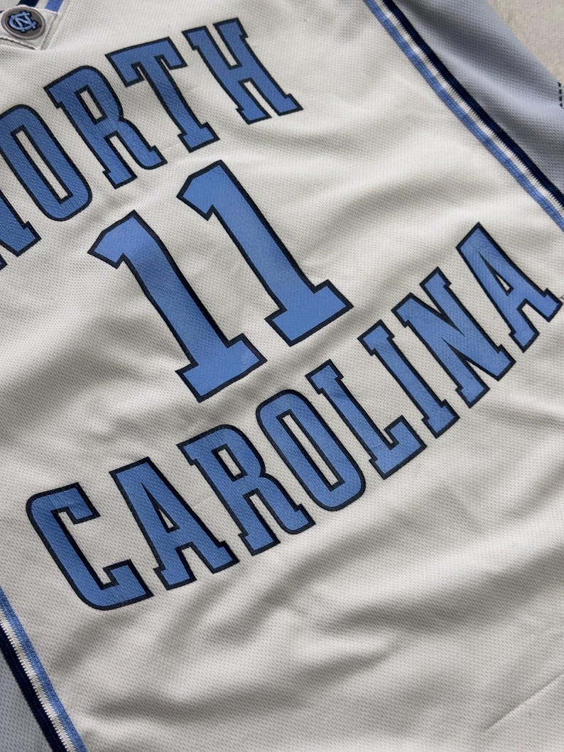 Op de afbeelding: Een lichtgrijs basketbalshirt met de tekst NORTH CAROLINA in blauwe letters met donkerblauwe omlijningen. Het nummer 11 staat in het midden gedrukt in hetzelfde blauwe en donkerblauwe kleurenschema. Een klein North Carolina logo zit op de kraag.