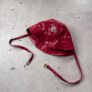 May include: A glossy dark red bucket hat with a shiny finish, featuring a circular Moncler logo patch on the front. The hat includes two long, matching red chin straps that hang down, each finished with a small gold-toned metal hardware detail.