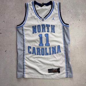Op de afbeelding: Een wit North Carolina basketbalshirt met lichtblauwe en marineblauwe biezen. De voorzijde bevat de tekst NORTH CAROLINA, het nummer 11, een Jumpman-logo en een Nike Elite-label. De zijkanten zijn lichtgrijs met een subtiel ruitpatroon.