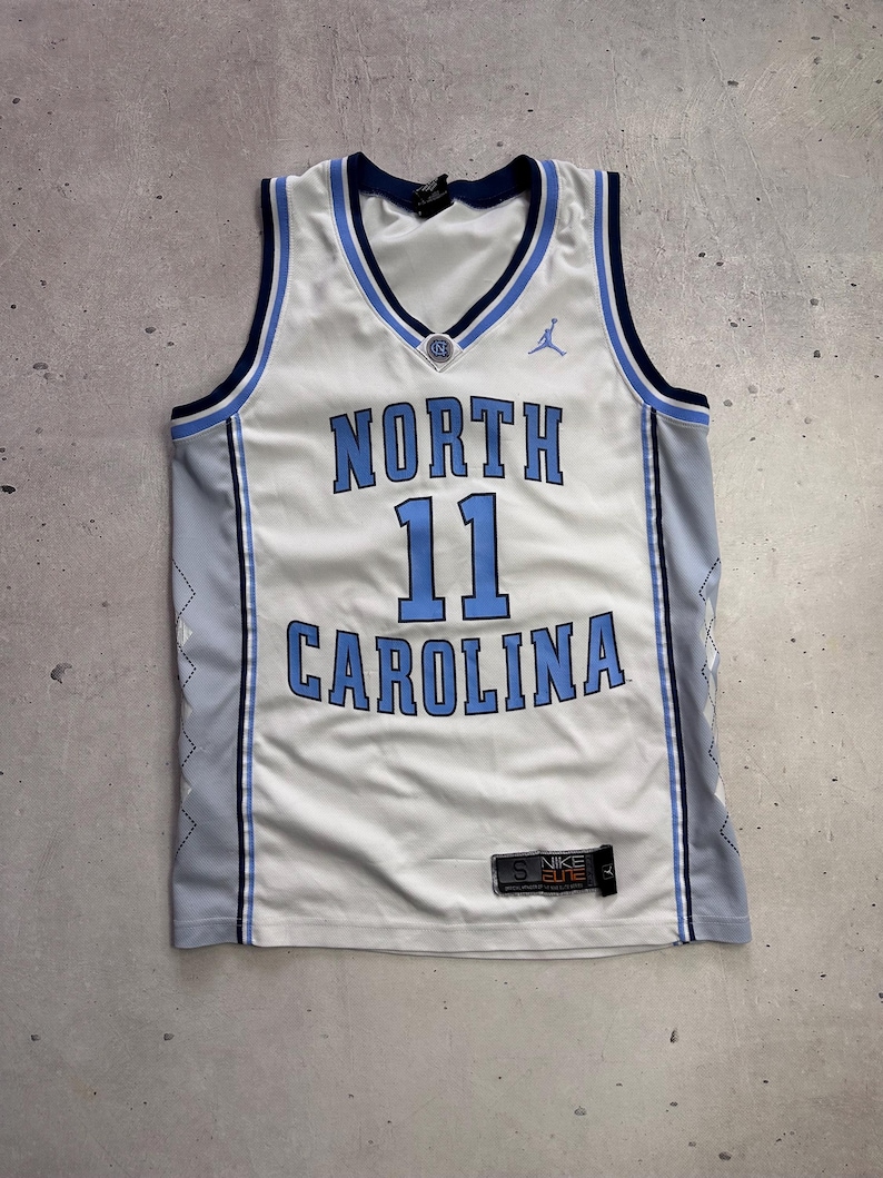 Op de afbeelding: Een wit North Carolina basketbalshirt met lichtblauwe en marineblauwe biezen. De voorzijde bevat de tekst NORTH CAROLINA, het nummer 11, een Jumpman-logo en een Nike Elite-label. De zijkanten zijn lichtgrijs met een subtiel ruitpatroon.