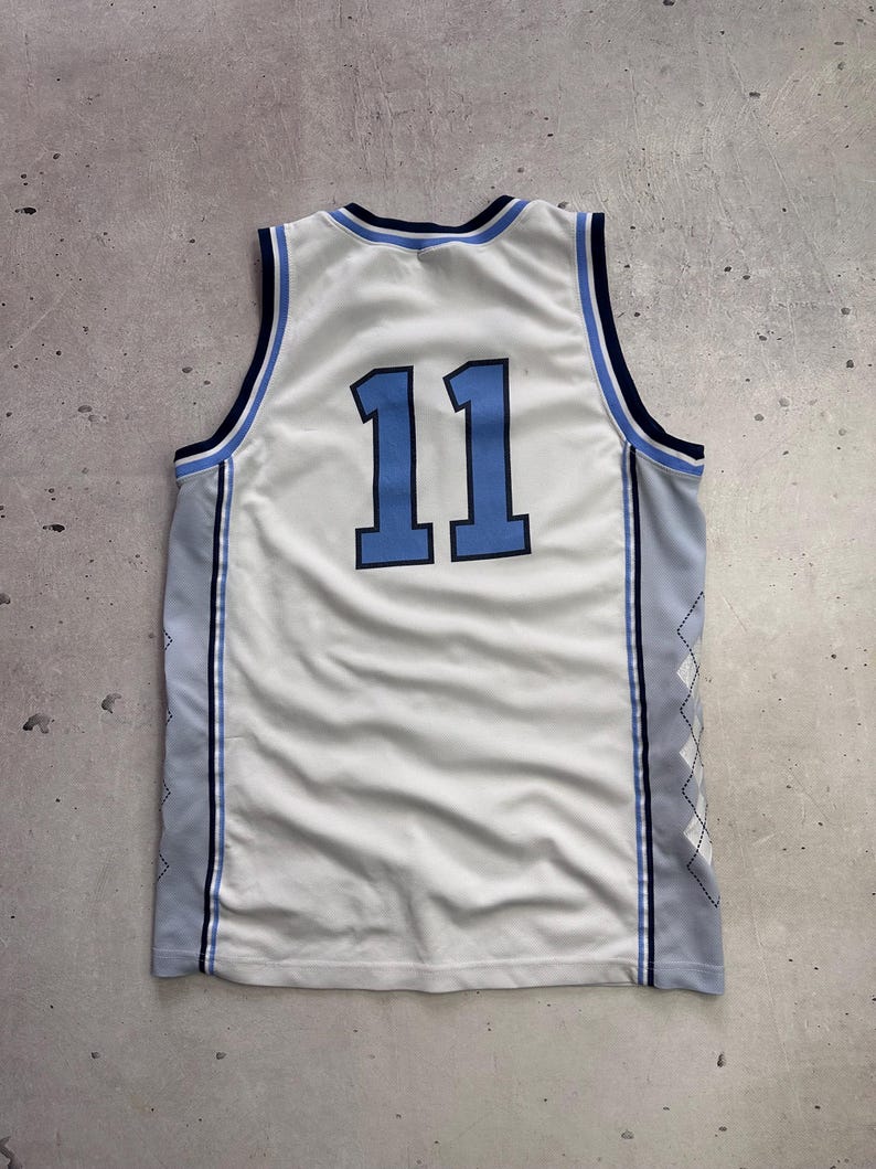 Op de afbeelding: Een wit mouwloos basketbalshirt met het nummer 11 in lichtblauw met een donkerblauwe omlijning. Het kledingstuk heeft donkerblauwe en lichtblauwe biezen bij de hals en armsgaten, met lichtgrijze zijpanelen voorzien van een wit ruitpatroon.