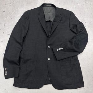 May include: A charcoal grey blazer featuring a notched lapel, two front buttons, and two large patch pockets. The sleeves are finished with four buttons at the cuffs and include dark elbow patches. The interior lining is a solid dark grey colour.