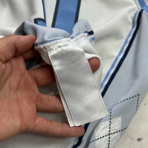 Op de afbeelding: Een wit sportshirt met lichtblauwe en donkerblauwe accenten, inclusief een groot nummer op de achterkant en een ruitpatroon aan de zijkant. Een wit stoffen waslabel met gedrukte tekst is bevestigd aan de binnennaad van het kledingstuk.