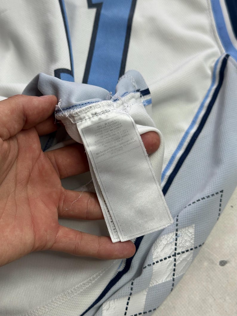 Op de afbeelding: Een wit sportshirt met lichtblauwe en donkerblauwe accenten, inclusief een groot nummer op de achterkant en een ruitpatroon aan de zijkant. Een wit stoffen waslabel met gedrukte tekst is bevestigd aan de binnennaad van het kledingstuk.
