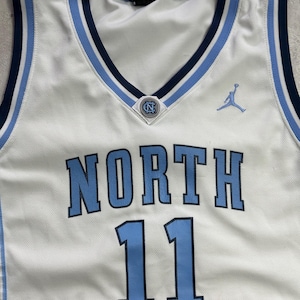 Op de afbeelding: Een wit basketbalshirt met lichtblauwe en marineblauwe biezen bij de V-hals en armsgaten. Op de voorzijde staat het woord NORTH in lichtblauw met marineblauwe omlijning, het nummer 11, een klein NC-logo en een lichtblauw Jumpman-logo.