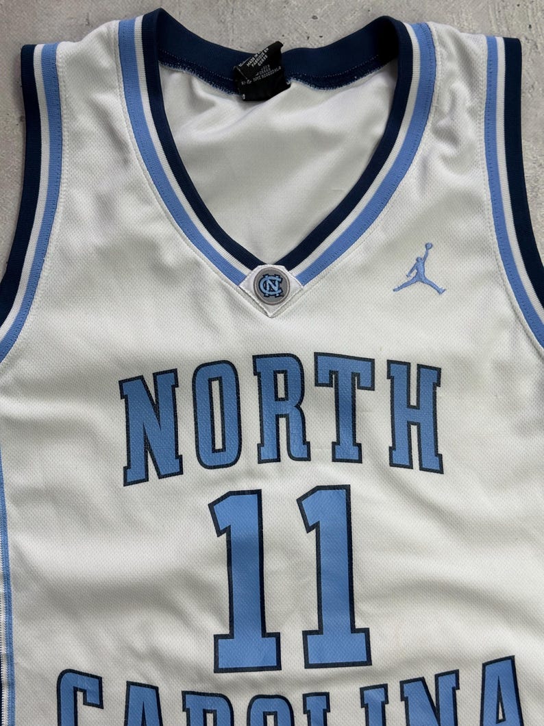 Op de afbeelding: Een wit basketbalshirt met lichtblauwe en marineblauwe biezen bij de V-hals en armsgaten. Op de voorzijde staat het woord NORTH in lichtblauw met marineblauwe omlijning, het nummer 11, een klein NC-logo en een lichtblauw Jumpman-logo.