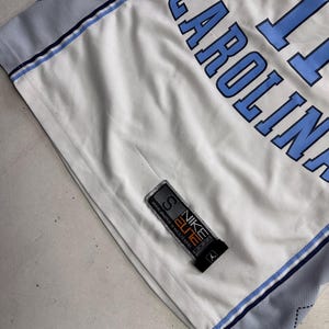 Op de afbeelding: Een wit basketbalshirt met lichtblauwe letters die CAROLINA en het nummer 11 spellen. Het shirt heeft lichtblauwe zijpanelen met een donkerblauw ruitpatroon en een zwart Nike Elite-label aan de onderkant met de maat S.