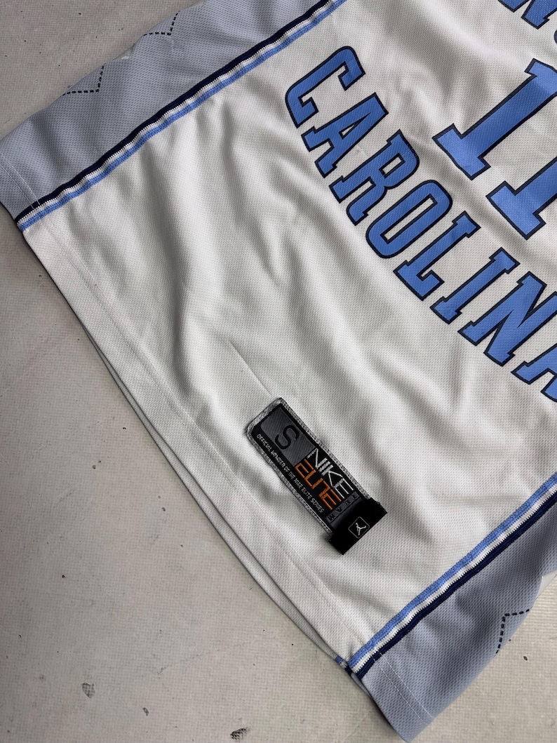 Op de afbeelding: Een wit basketbalshirt met lichtblauwe letters die CAROLINA en het nummer 11 spellen. Het shirt heeft lichtblauwe zijpanelen met een donkerblauw ruitpatroon en een zwart Nike Elite-label aan de onderkant met de maat S.