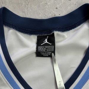 Op de afbeelding: Een wit shirt met een marineblauwe en lichtblauwe geribbelde V-hals. Een zwart label aan de binnenkant toont het Jordan Jumpman-logo, maat S, hoogte 173 en productiedetails waaronder Made in Korea. Een metalen schaarblad is zichtbaar.