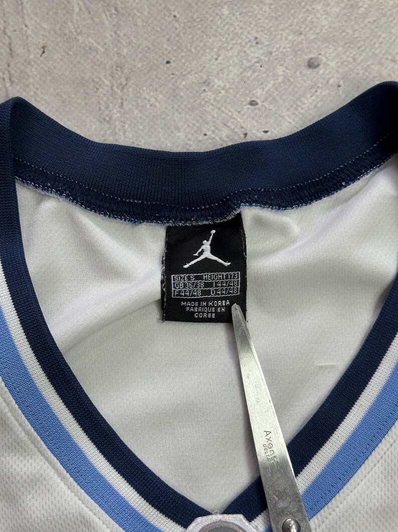 Op de afbeelding: Een wit shirt met een marineblauwe en lichtblauwe geribbelde V-hals. Een zwart label aan de binnenkant toont het Jordan Jumpman-logo, maat S, hoogte 173 en productiedetails waaronder Made in Korea. Een metalen schaarblad is zichtbaar.