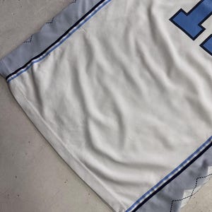 Op de afbeelding: Een wit sportshirt van mesh met lichtblauwe en donkerblauwe biezen aan de zijkanten. De zijpanelen hebben een subtiel grijs ruitpatroon met dunne zwarte stiksels. Een deel van een groot donkerblauw cijfer is zichtbaar aan de rechterkant.