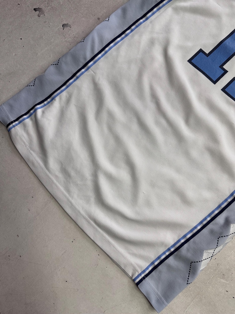 Op de afbeelding: Een wit sportshirt van mesh met lichtblauwe en donkerblauwe biezen aan de zijkanten. De zijpanelen hebben een subtiel grijs ruitpatroon met dunne zwarte stiksels. Een deel van een groot donkerblauw cijfer is zichtbaar aan de rechterkant.
