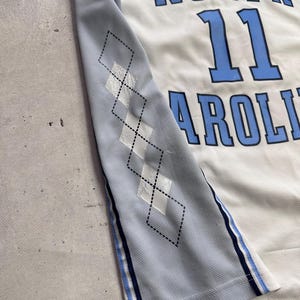 Op de afbeelding: Een wit en lichtblauw basketbalshirt met de tekst NORTH CAROLINA en het nummer 11 in blauwe letters. Het zijpaneel is lichtblauw met een zwart ruitpatroon en een gestreepte bies in marineblauw, wit en lichtblauw.