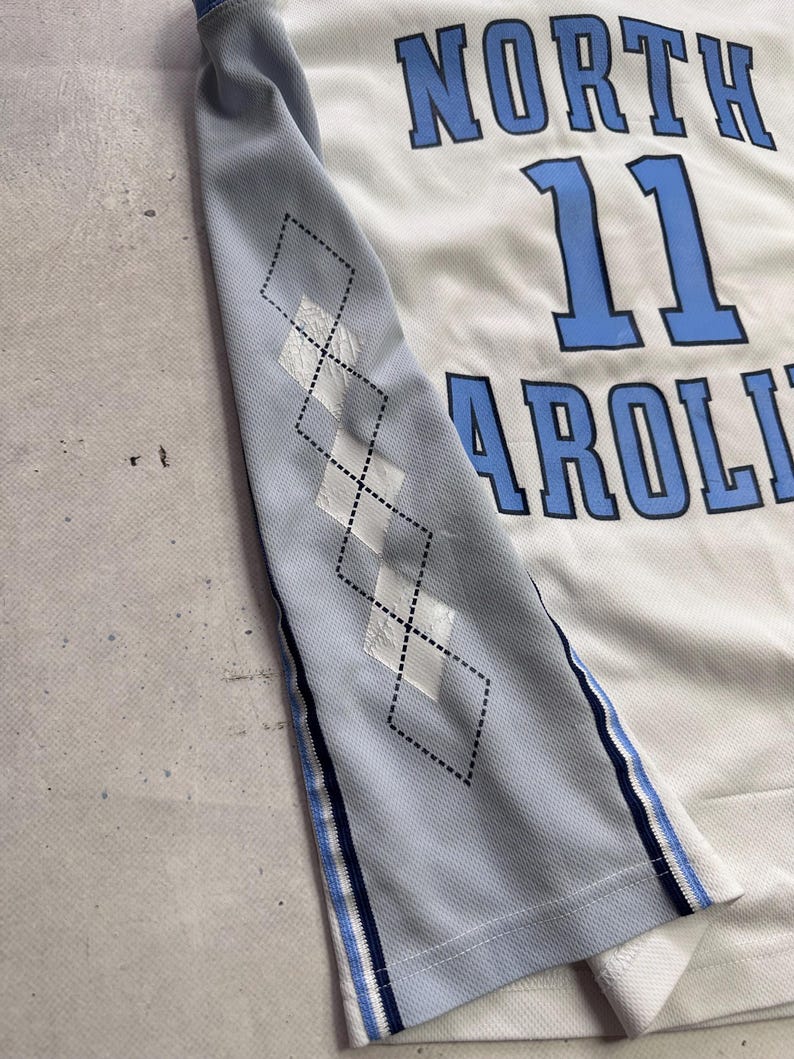 Op de afbeelding: Een wit en lichtblauw basketbalshirt met de tekst NORTH CAROLINA en het nummer 11 in blauwe letters. Het zijpaneel is lichtblauw met een zwart ruitpatroon en een gestreepte bies in marineblauw, wit en lichtblauw.