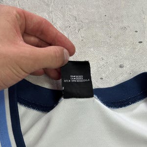 Op de afbeelding: Een wit kledingstuk met een donkerblauwe en lichtblauwe gestreepte kraag en een zwart binnenlabel met de tekst RN# 56323, CA# 05553 en RFC# NME 920220KL4. De stof oogt glad en lichtgewicht met een zichtbare geribbelde afwerking bij de hals.