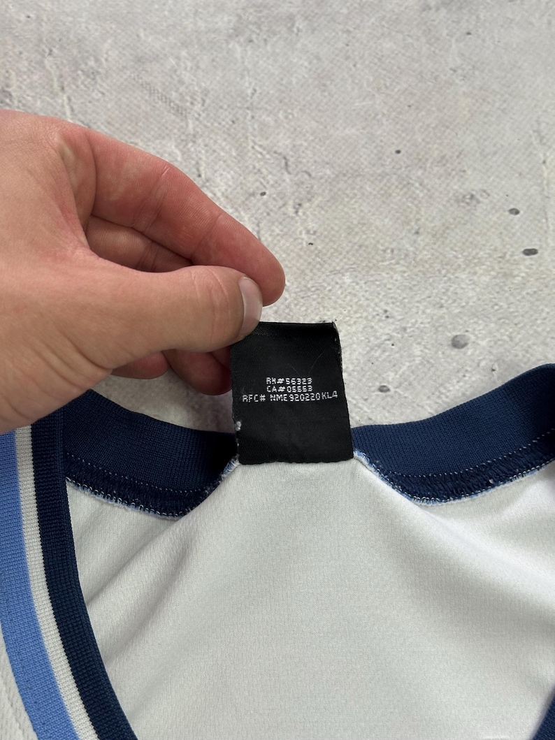Op de afbeelding: Een wit kledingstuk met een donkerblauwe en lichtblauwe gestreepte kraag en een zwart binnenlabel met de tekst RN# 56323, CA# 05553 en RFC# NME 920220KL4. De stof oogt glad en lichtgewicht met een zichtbare geribbelde afwerking bij de hals.