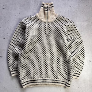 May include: A cream-colored knit sweater featuring a repeating black speckled pattern throughout the body and sleeves. The garment includes a high collar with a silver-toned metal zipper closure and black horizontal stripes on the cuffs and hem.