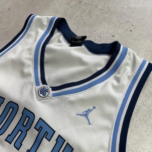 Op de afbeelding: Een wit basketbalshirt met lichtblauwe en marineblauwe biezen rond de V-hals. Het shirt toont het woord NORTH in lichtblauwe letters, een lichtblauw Jumpman-logo en een klein rond North Carolina-logo bij de halslijn.