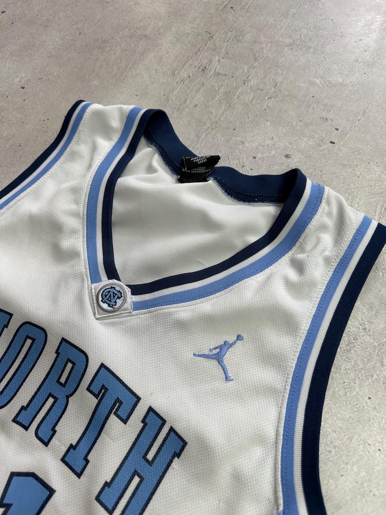 Op de afbeelding: Een wit basketbalshirt met lichtblauwe en marineblauwe biezen rond de V-hals. Het shirt toont het woord NORTH in lichtblauwe letters, een lichtblauw Jumpman-logo en een klein rond North Carolina-logo bij de halslijn.