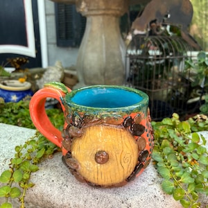 May include: A ceramic mug featuring a bright orange exterior with a prominent circular yellow door design on the front, surrounded by small dark brown stones. The handle is orange, and the interior of the mug is a vibrant, glossy turquoise blue.
