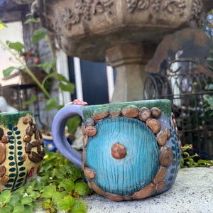 May include: A ceramic mug featuring a sculpted blue circular door with wood grain texture, surrounded by brown pebble-like accents. The mug has a light purple handle and a green textured rim. A small red and white mushroom sits atop the handle.
