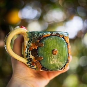 May include: A ceramic mug featuring a sculpted green circular door with a central brown knob, surrounded by textured brown and tan stone-like accents, a yellow handle, and blue leaf patterns painted on the side of the vessel.