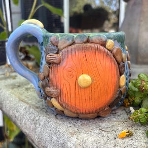 May include: A ceramic mug featuring a sculpted orange circular door with wood grain texture and a central knob. The exterior is decorated with small, multi-colored stones surrounding the door, set against a blue and green textured background.