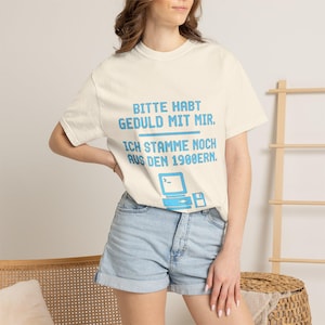 Könnte beinhalten: Ein cremefarbenes Kurzarm-T-Shirt mit blauem Text, der "BITTE HABT GEDULD MIT MIR. ICH STAMME NOCH AUS DEN 1900ERN." lautet, sowie einer Grafik eines alten Computers und einer Diskette unterhalb des Textes.