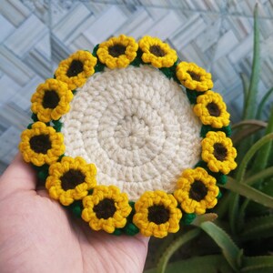 Peut inclure: Un dessous de verre rond en crochet de couleur crème, orné d'une bordure de onze petites fleurs de tournesol en relief avec des pétales jaune vif et des cœurs marron foncé, reliées par des accents de fil vert sur une base circulaire.