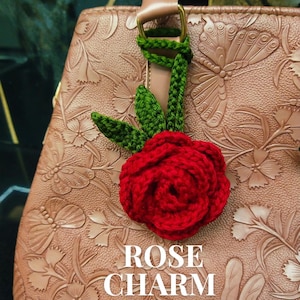 May include: A red crocheted rose flower charm with green leaves attached to a green crocheted strap. The charm is displayed on a tan leather handbag with an embossed butterfly and floral pattern. Text on the image reads ROSE CHARM Crochet Charm PDF Pattern.