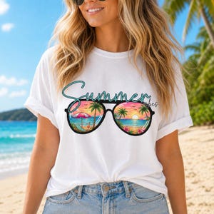 May include: A white short-sleeve t-shirt featuring a graphic of black sunglasses. The lenses display a colorful beach sunset scene with palm trees, a beach umbrella, and the text Summer vibes written in a teal cursive script above the glasses.