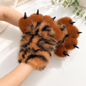 May include: Fingerless gloves made of soft, orange and black tiger-striped faux fur. The gloves feature plush, rounded paw pads in a solid orange color, each accented with a sharp, glossy black claw extending from the fingertip area.