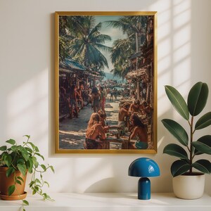 May include: A rectangular wall art print in a gold frame depicting a sunlit tropical beach scene with people sitting at outdoor tables under palm trees. The image shows a sandy path lined with rustic structures, lush green foliage, and figures.