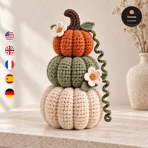 Puede incluir: Una decoración de calabazas de ganchillo apiladas que incluye tres calabazas en color naranja, verde salvia y crema. La calabaza superior naranja tiene un tallo marrón oscuro, y el conjunto está adornado con dos pequeñas flores blancas con centro amarillo y una larga enredadera verde.