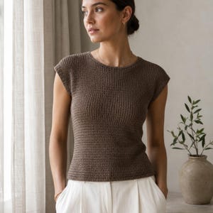 Puede incluir: Una blusa de punto marrón de manga corta con cuello redondo y un patrón de tejido texturizado. La prenda tiene una silueta ajustada con mangas cortas y se combina con pantalones blancos, creando una estética neutra y limpia.