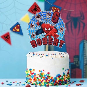 May include: A round cake topper featuring a red and blue superhero character in a web design, with the number 6 and the name ROBERT in red block letters. The topper is displayed on a white cake decorated with colorful sprinkles at the base.
