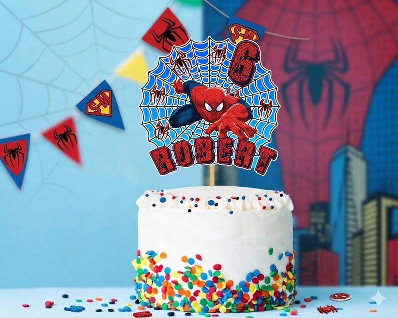 May include: A round cake topper featuring a red and blue superhero character in a web design, with the number 6 and the name ROBERT in red block letters. The topper is displayed on a white cake decorated with colorful sprinkles at the base.